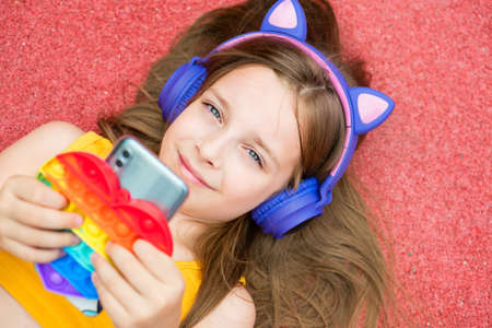 Pretty School girl hold smartphone and Anti-stress colorful toy pop . Young girl chatting friends, listen audio in a music app, modern technology earphones.の写真素材