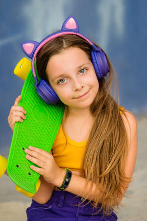Portrait young hipster little girl with Skateboard and trendy wireless headphones on outdoor. Cute primary school student posing with longboardの写真素材