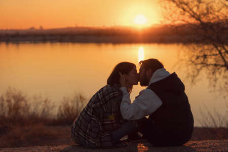 Romantic loving couple kissing while sitting at sunset by lake. Lovers spend time together outdoors. Authentic real people in casual style. Lifestyle Candid portrait. .の写真素材