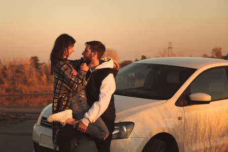Man and woman in holiday honeymoon trip. Couple is hugging in twilight near car on the road outdoors. Lovers traveling together. Freedom love lifestyle. Romantic sunset journeyの写真素材