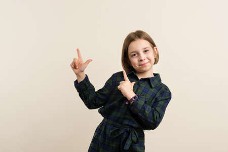 Cute Little Girl 9s in plaid shirt dress points two fingers in beige background. Child pointing index fingers copy space for ad. Wow, look, advertise here.の写真素材