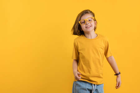 Joyful Little Kid Girl 8-10 Years Old in Yellow T-shirt and Eyeglasses on Daffodil Color Trend Wall Background Studio Portrait. Gender Neutral Child Having Fun. Childhood Lifestyle Concept.の写真素材