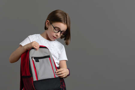 Smart Little Girl 9s Wearing school uniform and in stylish round glasses with book bag on grey...
