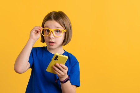 Little Girl 9s in Basic Blue T-shirt and Yellow Glasses Using Smartphone Scrolling and Reading News. Child holding cell phone with Amazed Look on Yellow Background. Children and Technology. Copy spaceの写真素材