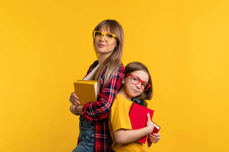 Young mom and funny daughter in casual clothes standing back to back and hugging textbooks on yellow background studio. Family having fun together. Educationの写真素材