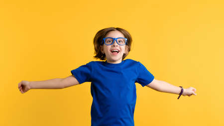 Happy Little Girl 8-10 Years Old in Blue T-shirt and Glasses Jumping and Having Fun on Yellow Studio Background. Child Playful, Laughing, Smiling. Childhood, Emotions, Facial Expression.の写真素材
