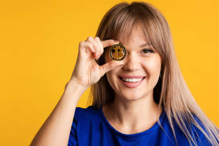 Smiling Girl in Blue Wear Holds Golden Physical Bitcoin in Front her Eye. Closeup fun emotional...