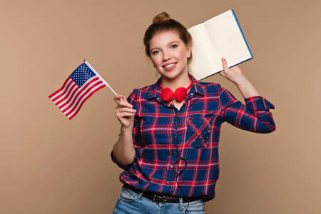 Young Woman in Casual Shirt and headphones with american flag and open notebook indoor beige background. Smiling exchange student girl looking at camera. Study abroad concept.の写真素材