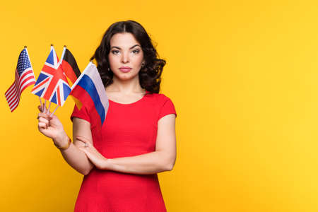 Female with set of flags of different countries of the world looking at camera. Serious and confident Young woman wearing red dress on yellow background. Political Union and unification conceptの写真素材