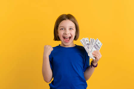 Child Holding Wad of Dollar Bills and Smiling. Fortunate Little Girl 8-10 Years Old in Basic Blue T-shirt Doing Winner Gesture on Yellow Background. Children Financial Literacy Concept.の写真素材