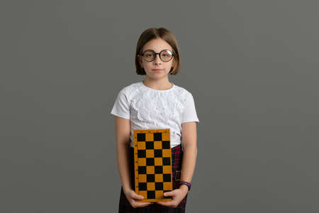 Calm and Successful Little Girl 9s Wearing School Uniform Holding Chessboard on gray background. Learn to play chess. Child chess player.の写真素材