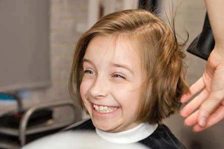 child sitting in hairdresser chair delightedly rejoices at new bob haircut. Hairdresser dries wet hair after cutting with hairdryer. Hair styling process in barbershop. Happy little girlの写真素材