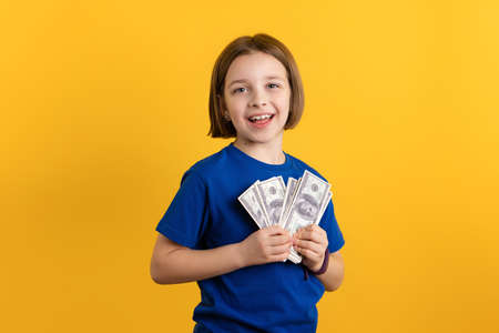 Successful Little Girl 8-10 Years Old in Basic Blue T-shirt on Yellow Background. Child Holding Dollar Banknotes and Smiling. Children Financial Literacy Concept. Invest in Futureの写真素材
