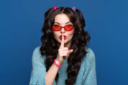 Funny girl with two ponytails put index finger to lips on blue background. Young woman wears in blue...