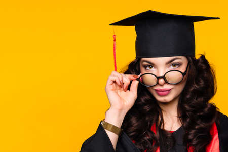 Close up self-confident portrait young woman graduate in graduation hat and eyewear on yellow wall. Female best student wearing graduation cap and ceremony robe holding glasses and looks at camera.の写真素材