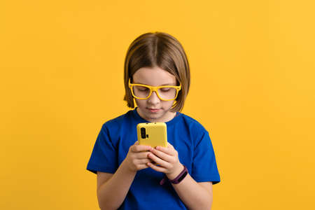 Girl 9s in Basic Blue T-shirt and Yellow Glasses Using Smartphone Scrolling and Reading News. Child Types Text Message Enjoys Online Communication on Yellow Background. Children and Technologyの写真素材