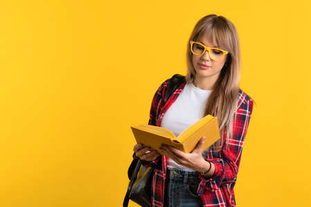 Smart student girl in basic clothes and yellow glasses reads paper book in studio on yellow background. Education concept.の写真素材