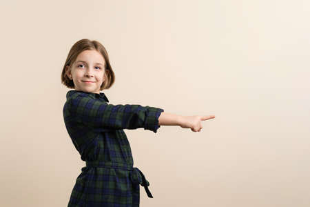 Joyful Little Girl 9s in Plaid Shirt Dress Points Index Finger in ivory Background. Smart Child Have Excellent Idea. Showing promo. Inviting use link. Give direction.の写真素材