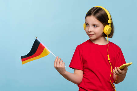 Girl 9-11 y.o. in basic red t-shirt and yellow headphones with cellphone looking away. Child holding German flag and smartphone. Languages courses online. Learning Deutsch language. Listeningの写真素材