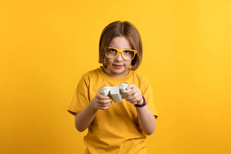 Child with gamepad playing video game. Little Girl 8-10 Years Old in Yellow T-shirt and Eyeglasses...