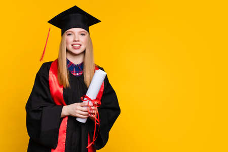 Portrait girl graduate with graduation hat and diploma on yellow background. Blonde young woman wearing graduation cap and ceremony robe holding Certificate tied with red ribbon. Education Conceptの写真素材