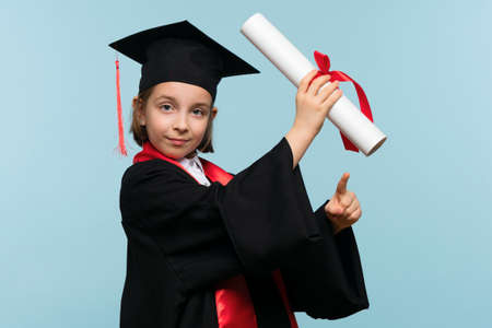 Graduate celebrating graduation. Child Pointing on Certificate Diploma. Whizz kid girl wearing graduation cap and ceremony robe on light blue background looking at camera. Education Concept.の写真素材