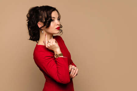 Side view young woman in red dress and gold watch pointing with index finger at pearl earring in...