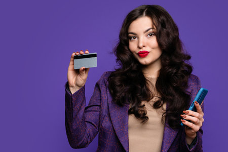 Elegant woman in business suit holds plastic credit card and smartphone on purple background indoor. Cashless, contactless payment, money transfer and people concept. Modern digital technologyの写真素材