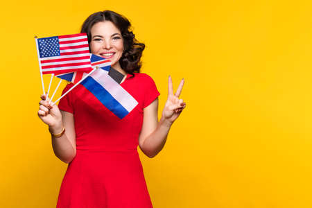 Woman in red dress shows bunch of diverse flags cheerfully smiling at camera over yellow studio background. Smiling girl shows V-sign, winner gesture and peace sign. Female recommends language schoolの写真素材