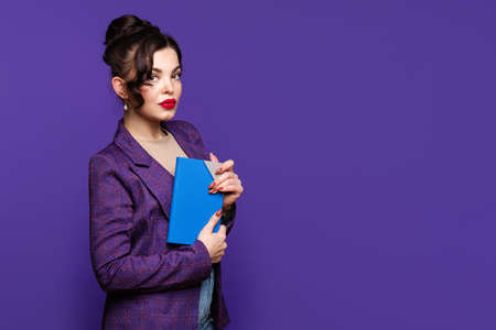 Focused young businesswoman in purple jacket posing on purple background. Woman holds planner,...