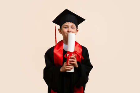 Whizz kid 9-11 year wearing graduation cap and ceremony robe Kissing Paper Certificate of Education. Elementary school graduate boy with certificate diploma tied with red ribbon in studio.の写真素材