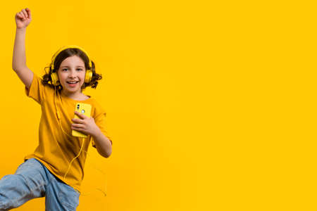 Cheerful short curly hairdo school girl dance to favorite song moves to rhythm of music holds modern smartphone expresses happiness and joy.の写真素材