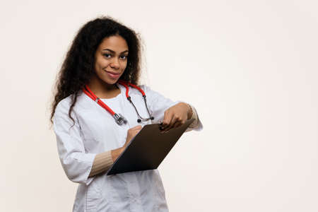 African American Female Pediatrician with Digital Tablet and Happy Girl Patient on Light background. Friendly Dark Skinned Woman Doctor in Uniform Talking to Patient. Child Holds Soft Toy in Handsの写真素材