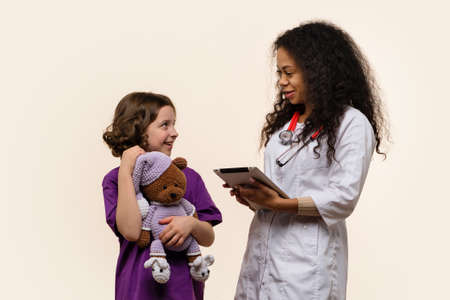 African American Female Pediatrician with Digital Tablet and Happy Girl Patient on Light background. Friendly Dark Skinned Woman Doctor in Uniform Talking to Patient. Child Holds Soft Toy in Handsの写真素材