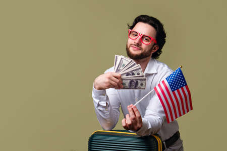 Bearded Man Traveler Tourist holds Fan Cash Dollars Banknotes and Flag USA. Guy in Casual Outfit and...