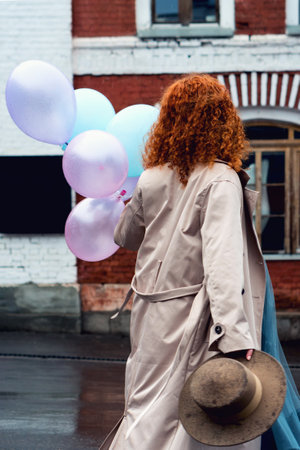 Stylish Portrait Curly Red Haired Woman in Makeup Room Near Mirror. Gorgeous Girl in Shiny Evening Dress and Black Jacket going to Fix Make up in Dress Room. Female Before Going to Partyの写真素材