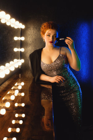 Stylish Portrait Curly Red Haired Woman in Makeup Room Near Mirror. Gorgeous Girl in Shiny Evening...