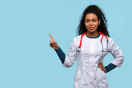 Friendly African American Female Physician Medical Advisor Shows Heart Gesture with Two Hands on Blue Studio Wall. Smiling Woman Doctor GP Wears Hospital Uniform with Phonendoscope. health careの写真素材