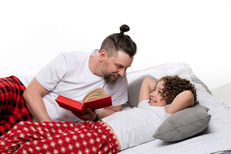 Family in plaid sleepwear sit on bed and read tea leaves on white studio background. Dad and curly short-haired daughter in pajamas. Daddy and female child girl spend time together for morning coffeeの写真素材