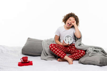 Curly short-haired girl in red plaid pajama pants and t-shirt holds large alarm clock. Sleepy female child rubbing her eyes in white background studio. Preteen relaxing at home. Wake up earlyの写真素材
