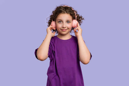 Side view curly short-haired girl 10-12s wears purple basic t-shirt and pink wireless headphones. Female child listening to music, podcast, audiobook on lilac studio background. banner advertisingの写真素材