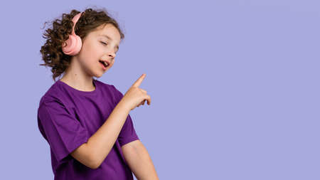 Side view curly short-haired girl 10-12s wears purple basic t-shirt and pink wireless headphones. Female child listening to music, podcast, audiobook on lilac studio background. banner advertisingの写真素材