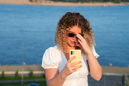 Attractive curly blonde girl talking on cell phone. Young caucasian woman at sunset summertime and sea outdoor background. Lifestyle leisure. city life conceptの写真素材