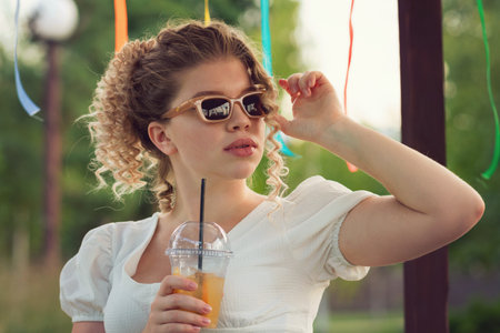 Attractive blonde curly girl in sunglasses and summer outfit enjoying lemonade, non-alcoholic cocktail at party in city park on open veranda. Cute young woman drinking. summer vacationの写真素材