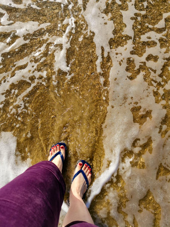 Female legs wearing flip-flops on the beach covered with sea foam. top view. beach accessoriesの写真素材