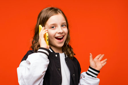 Teenager girl happily and emotionally talks on phone and looks at camera. Schoolgirl in bomber with...