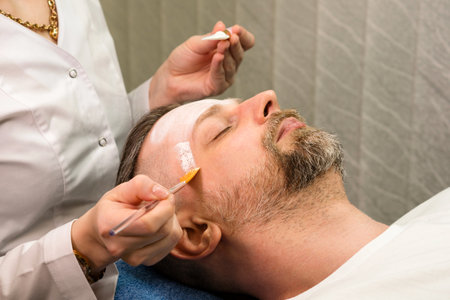 Cosmetologist applying cosmetic mask on man's face in spa salon. Handsome bearded man getting facial skin care treatment.の写真素材