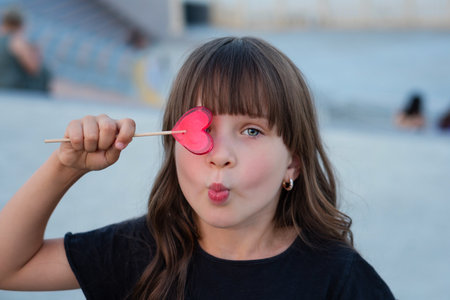 Funny little girl posing outdoors with red heart shaped lollipop. The child closed one eye with candy and makes grimacesの写真素材