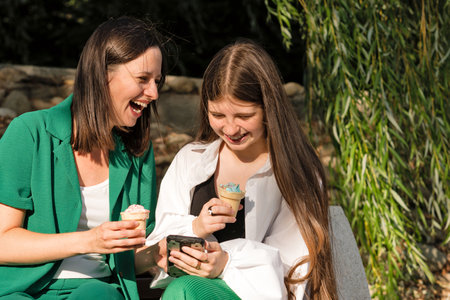 Cheerful and happy mom and teenage daughter eat ice cream on walk in park in summer on sunny day....