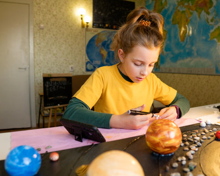 Cheerful girl engrossed in painting her mini planets, surrounded by classroom ambiance and a teddy...